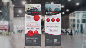Two National Quantum Office (NQO) X-stand banners erected in a modern exhibition hall with blurred background lights and booths. Left banner: Diverse youth group photo, red circular icons, structured text sections, QR code. Right banner: "Building a Quantum Future Together" headline, heart and blood drop icons, partner logos in blue circles, QR code. Red and gray design scheme creates immersive environmental branding for quantum research showcase.